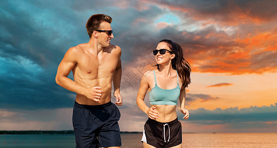 健身,运动生活方式的幸福的夫妇穿着运动服装太阳镜跑海上日落天空的背景穿着运动服的夫妇海滩上跑步图片