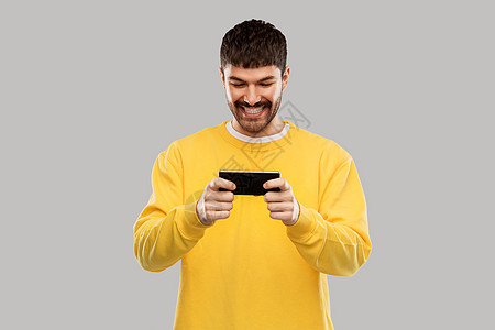 技术,沟通人的快乐的微笑年轻人智能手机上玩游戏灰色背景带着智能手机的快乐微笑的年轻人图片