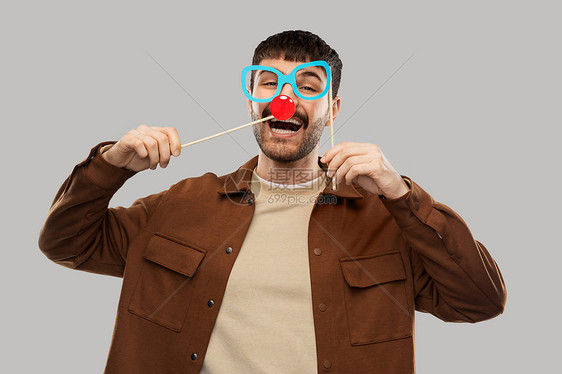 派道具,照片亭人们的快乐的微笑年轻人带着纸眼镜红色小丑鼻子灰色背景带着眼镜红色小丑鼻子的快乐微笑的男人图片