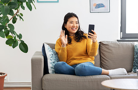 人,技术沟通快乐微笑的亚洲年轻妇女穿着黄色毛衣坐沙发上,家里用智能手机打电话有智能手机的女人家有视频通话图片