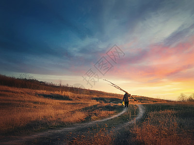 老人的身影走条乡间道路上,肩上扛着干枯的树枝,夕阳的天空背景下田园乡村景观,深秋季节泥土轨道穿过金色的干草图片