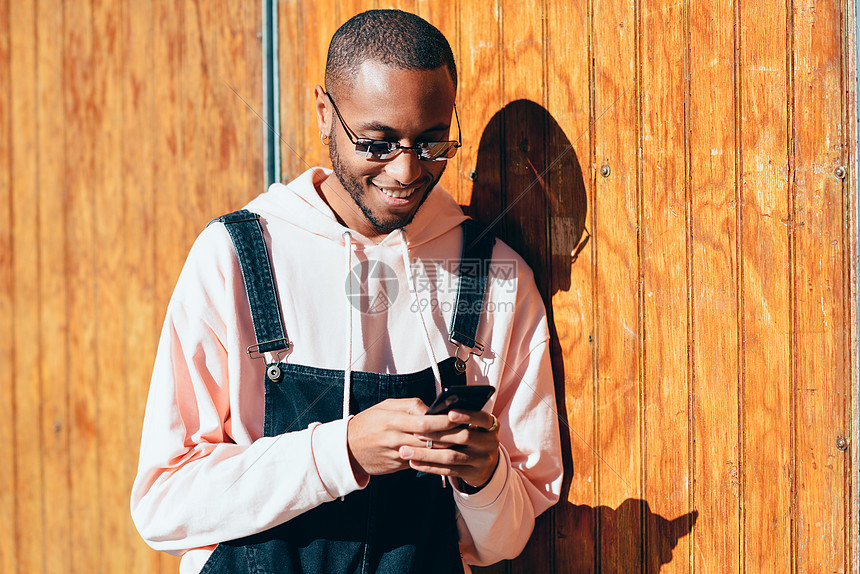 年轻的黑人男子穿着休闲服装太阳镜,木制背景下用智能手机千禧年的非洲人带着围嘴裤户外微笑图片