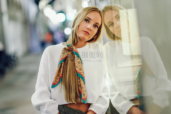 金发女孩穿着白色毛衣站街上,背景上有分散注意力的城市灯光晚上梳着辫子的漂亮女人反映商店的橱窗里图片