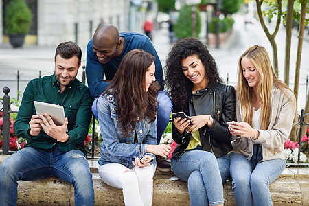 城市背景下用智能手机平板电脑的多民族年轻人群体女人男人街上笑着,穿着便装图片