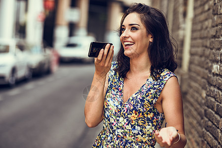 微笑的年轻女人户外用她的智能手机录制语音音符城市背景下穿花裙的女孩技术微笑的年轻女人她的智能手机上录制语音音图片