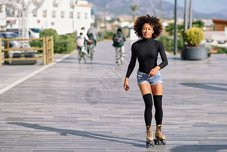 年轻合身的黑人妇女溜冰鞋骑户外的城市街道上阳光明媚的日子里,微笑的女孩留着AFRO的发型人们背景下徘徊骑着溜冰鞋背景图片