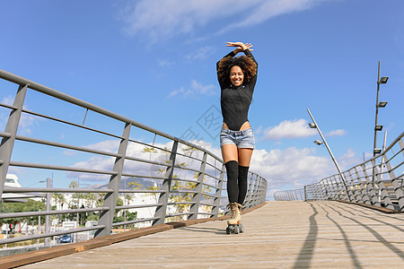 黑色女人,AFRO发型,穿着溜冰鞋,户外骑着城市桥,张开双臂阳光明媚的日子里,微笑的年轻女溜冰天空丽的云彩阿夫图片