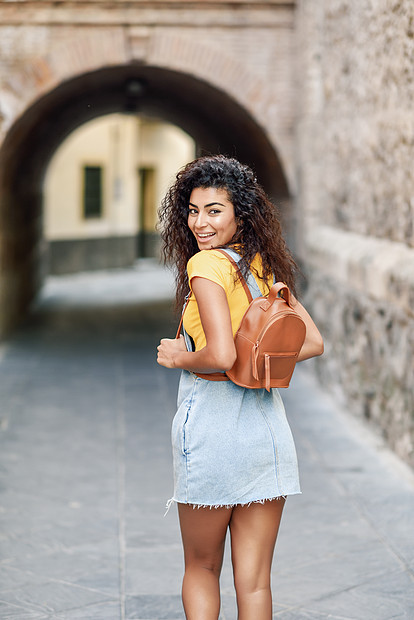 带背包的年轻阿拉伯女人户外的后视镜旅行者女孩穿着休闲服街上快乐的女穿着黄色T恤城市背景的牛仔连衣裙年轻的北非女图片