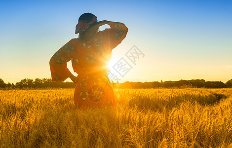 非洲妇女穿着传统的衣服,站着,看着,手着眼睛,大麦或小麦作物的田野日落或日出图片