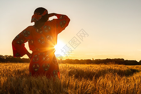 非洲妇女穿着传统的衣服,站着,看着,手着眼睛,大麦或小麦作物的田野日落或日出图片