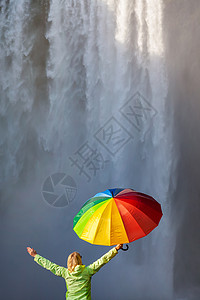 轻的金发女人穿着件绿色的外套夹克手臂举着庆祝,瀑布前着把五颜六色的雨伞图片