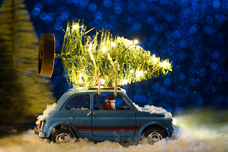 圣诞老人穿着蓝色复古玩具车,节日背景上送圣诞或新年照明树圣诞老人送圣诞或新年树背景图片