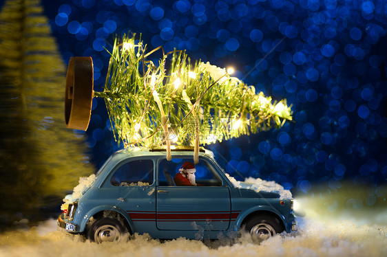 圣诞老人穿着蓝色复古玩具车,节日背景上送圣诞或新年照明树圣诞老人送圣诞或新年树图片
