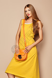 穿着黄色连衣裙的时尚年轻女人的肖像时尚的春季夏季服装的女模特米色背景上摆姿势的女孩时尚的发型带橙色手提包的金图片