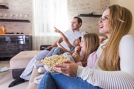 美丽的年轻父母他们的孩子家里坐沙发上看电视吃爆米花微笑父母孩子看电视图片