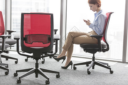 年轻的女商人坐办公室的红色座位上,智能手机上发短信图片