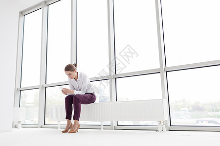 年轻的女商人新空办公室坐暖气片上用智能手机图片