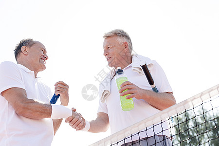 阳光明媚的日子里,微笑的男人网球场上站着,着晴朗的天空,边握手,边低角度观看图片