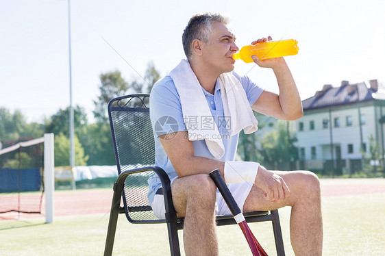 疲惫的成熟男人阳光明媚的日子里坐网球场的椅子上喝着瓶子图片