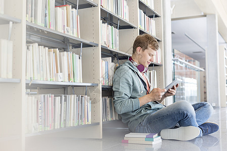 学生图书馆坐地板上时,用数字平板电脑着书架图片