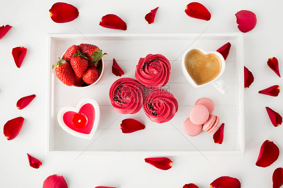 情人节糖果纸杯蛋糕与红色奶油霜,马卡龙,心形咖啡杯,蜡烛草莓托盘与玫瑰花瓣托盘上的款待情人节图片