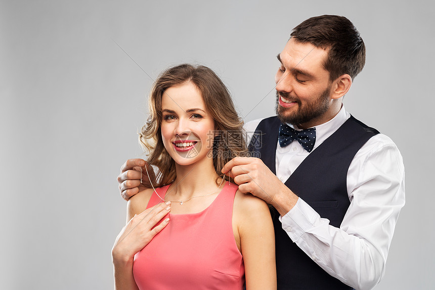 夫妇,珠宝人的快乐的男人把项链与钻石吊坠他的女朋友灰色背景快乐的男人把项链戴女朋友身上图片