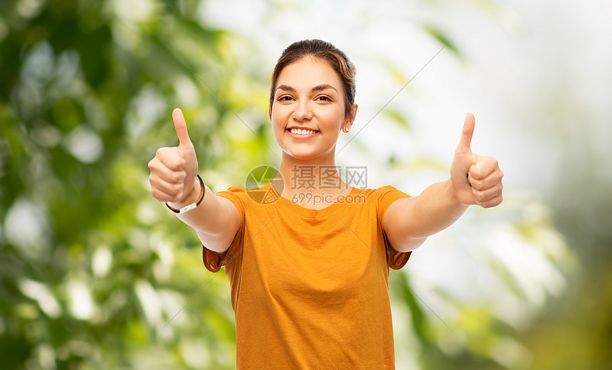 手势人的快乐微笑的轻女人十几岁的女孩穿着橙色的t恤,绿色的自然背景上竖大拇指十几岁的女孩自然的背上竖大拇图片