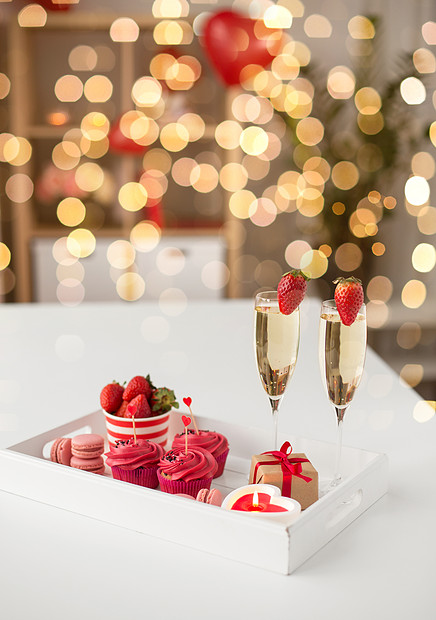 情人节糖果纸杯蛋糕与红色霜心形鸡尾酒棒,香槟杯与草莓,蜡烛礼品盒托盘上的节日灯情人节的红色糖果图片