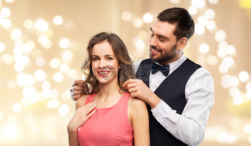 夫妇,珠宝人的快乐的男人把项链与钻石吊坠他的女朋友米色背景节日灯快乐的男人把项链戴女朋友身上图片