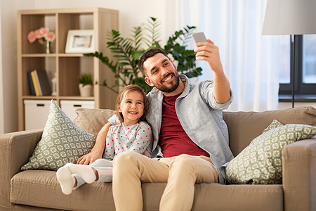 人,家庭技术快乐的父亲小女儿坐家里的沙发上用智能手机自拍父亲女儿家自拍图片
