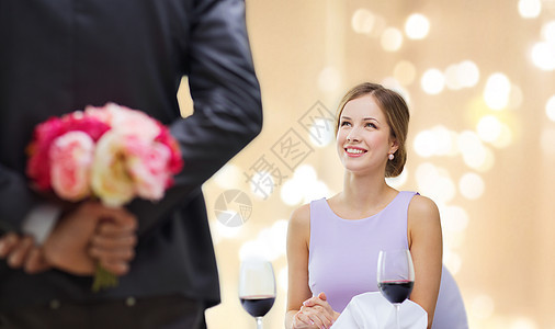 情侣,约会情人节微笑的轻女人看着男人背后的花餐厅的节日灯米色背景轻的女人看着着花的男人图片