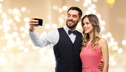 技术人们的快乐的夫妇穿着派服装,用智能手机米色背景下用节日灯自拍幸福的夫妇用智能手机自拍图片