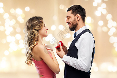 爱,夫妇,求婚人的快乐的男人给戴着节日灯的米色背景的女人小红盒子里的钻石订婚戒指男人情人节给女人订婚戒指图片