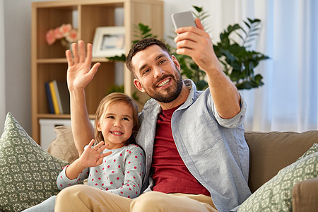 人,家庭技术快乐的父亲小女儿坐家里的沙发上用智能手机自拍父亲女儿家自拍图片