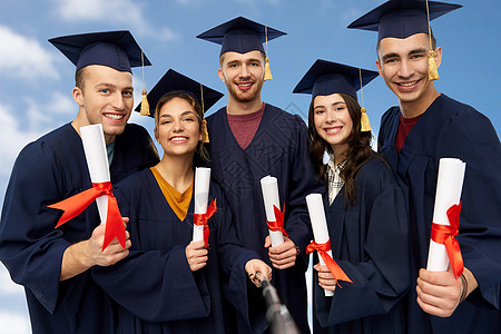 教育毕业人的群快乐的研究生穿着灰浆板学士学位礼服,用Slfie棒蓝天云层背景上拍照毕业证书自拍的快乐毕业生图片