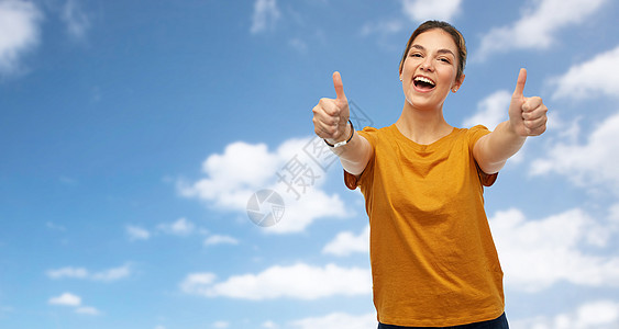 手势人的快乐微笑的轻女人十几岁的女孩穿着橙色的t恤,蓝天云彩的背景上竖大拇指穿着T恤的女人十几岁的女孩竖图片