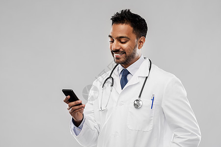 医学,技术医疗微笑的印度男医生穿着白色外套,智能手机听诊器灰色背景微笑的印度男医生与智能手机图片