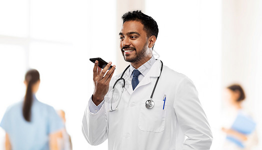 医学,技术医疗微笑的印度男医生穿着白色外套,带听诊器,医院背景下用语音命令记录器微笑的印度男医生打电话给智能图片