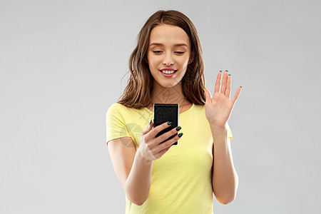 技术人的微笑的轻妇女十几岁的女孩穿着空白的黄色T恤,视频呼叫智能手机挥动手灰色背景微笑的十几岁女孩视频呼图片