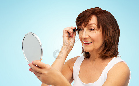 美丽,化妆老人的微笑的老年女子与镜子涂睫毛膏睫毛蓝色背景微笑的老妇女用镜子涂睫毛膏图片