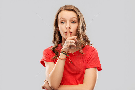 沉默,秘密人的十几岁的女孩穿着红色T恤,用手指灰色背景上的嘴唇上着沉默的手势十几岁的女孩穿着红色T恤,嘴唇上手图片
