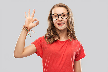 教育,学校人的微笑的十几岁的学生女孩戴着眼镜红色T恤,灰色背景上OK手牌微笑的学生女孩戴着眼镜,出良好的图片