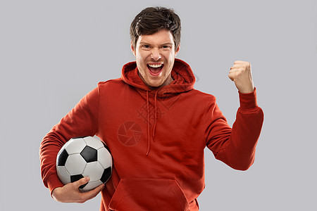 运动,休闲游戏获胜快乐的人足球迷穿着红色帽衫,带着足球庆祝胜利,并灰色背景上拳泵手势快乐的人足球迷,足球获胜图片