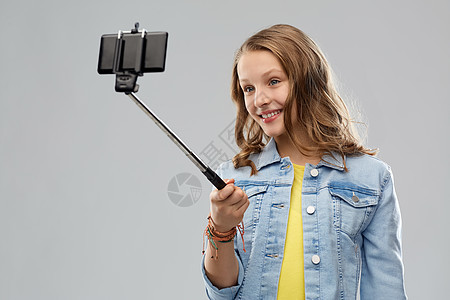 技术人们的快乐的微笑少女用智能手机灰色背景上的自拍杆上拍照十几岁的女孩用智能手机自拍图片