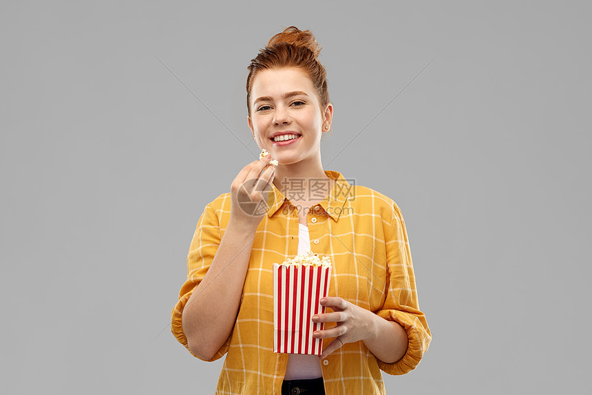 快餐人们的微笑的红色头发十几岁的女孩穿着格子衬衫,吃爆米花条纹桶灰色背景微笑的红头发少女吃爆米花图片