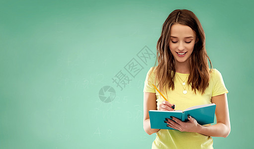 教育,学校,灵感人的微笑的十几岁的学生女孩穿着黄色T恤,用铅笔绿色粉笔板背景上写日记笔记本学生女孩绿色的笔记本图片