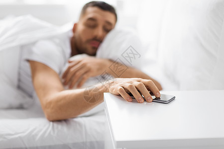 技术人的困倦的轻人伸手床头柜上的智能手机家里的床上困倦的轻人床上伸手智能手机图片