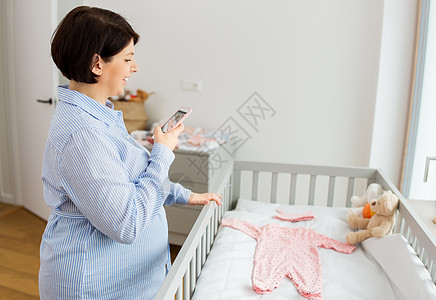 母亲,怀孕托儿所的快乐的怀孕中妇女与智能手机拍照婴儿衣服床上家孕妇给婴儿衣服拍照图片