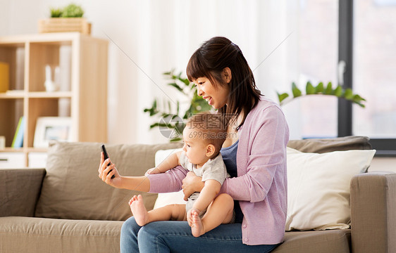 家庭母亲的快乐微笑的轻亚洲母亲与小婴儿儿子家里用智能手机自拍亚洲母亲带着小儿子家自拍图片