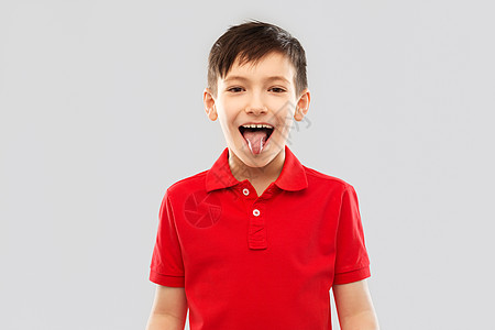 童,表情趣的快乐的小男孩穿着红色马球T恤,灰色背景上展示他的舌头穿着红色马球T恤的男孩露出舌头图片